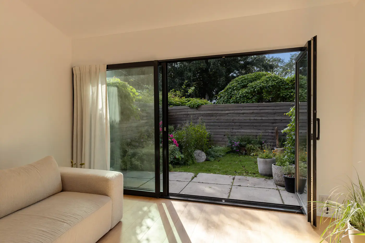 Woonkamer met een brede zwarte aluminium schuifpui die openstaat naar een kleine tuin